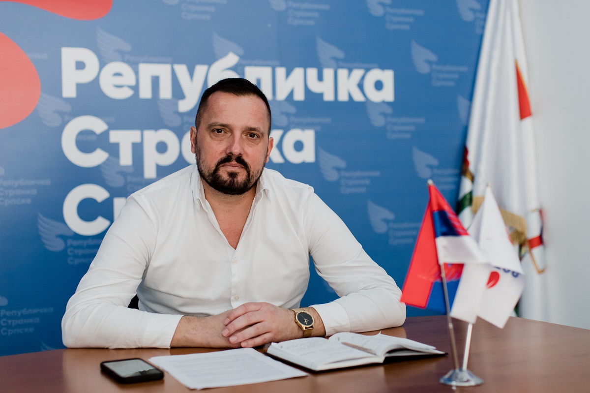 Muskarac sjedi za stolom i gleda pravo u kameru.